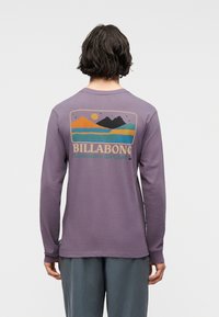 Άτομο με κοντά σκούρα μαλλιά φοράει μωβ μπλούζα με μακριά μανίκια που έχει ένα πολύχρωμο γραφικό βουνού και ήλιου με το κείμενο "BILLABONG Adventure division" στην πλάτη.