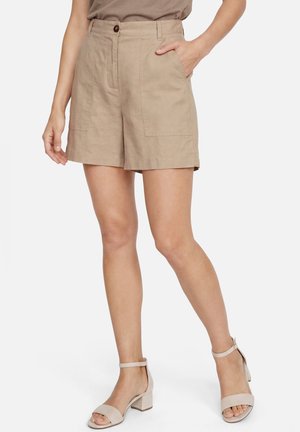 Femme portant un short beige taille haute avec de grandes poches devant et une fermeture à boutons, associé à des sandales beiges à talons blocs avec bride à la cheville.