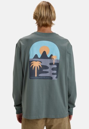 Vaaleatukkainen henkilö pukeutuu vihreään pitkähihaiseen paitaan, jonka selässä on auringonlaskun, vuorien ja palmupuiden kuvio.