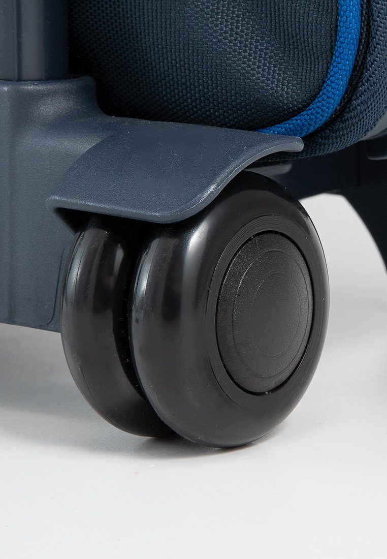 Esquina de una maleta rodante azul oscuro con un rodillo doble negro. Tejido texturizado visible a lo largo del cuerpo de la maleta. Componentes de plástico duradero.