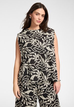 Frau trägt ärmelloses schwarzes Oberteil und Hose mit passenden elfenbeinfarbenen abstrakten Blattmustern, steht vor einem schlichten hellen Hintergrund.