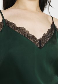 Caraco en soie verte avec bordure en dentelle noire, équipé de bretelles réglables et d'un décolleté en V. Texture lisse avec des accents délicats en dentelle.