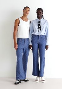 Două modele stau împreună, purtând blugi albaștri cu un model. Una poartă un top alb fără mâneci, iar cealaltă o cămașă albastră deschisă cu un accent de cravată neagră.