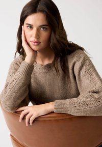 Brązowy sweter z dzianiny o teksturowanym wzorze, z falbankowym dekoltem i ściągaczami na mankietach. Model trzyma rękę na brązowym krześle.
