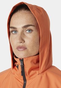 Oranje waterdichte jas met een hoge kraag en capuchon, voorzien van een frontale rits en textiel met een structuur. Accenten zijn zwarte hardware.