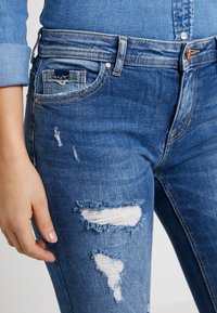 Jeans de denim azul con un diseño ajustado, parches desgastados con bordes deshilachados y un estilo clásico de cinco bolsillos.
