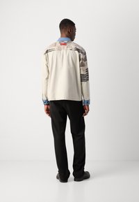 Hellbeige Hemd mit gemusterten Ärmeln, die gemischte Grafiken zeigen, Denim-Kragen und schwarze Hose. Modell von hinten, stehend vor einem neutralen Hintergrund.