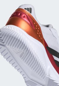 Witte sneaker met een gestructureerd bovenwerk, oranje en rode accenten op de hiel en zwarte strepen aan de zijkanten. Dikke, wit met patroon versierde zool.