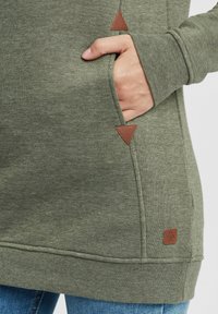 Olivgrön sweatshirt med en texturerad tyg, med sidofickor, bruna triangulära detaljer och en liten logopatch vid fållen.
