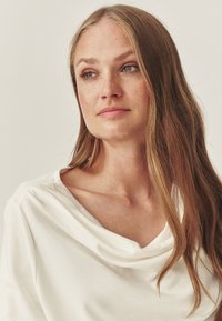 Hvid cowl-neck top lavet af blødt stof, med en afslappet pasform. Lange, lige hår falder ned over skuldrene og fremhæver udskæringen.