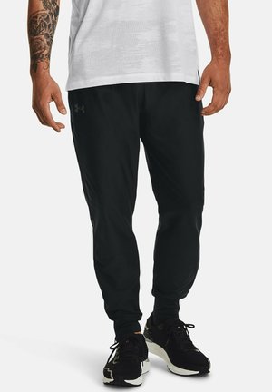 Schwarze Sporthosen aus leichtem Material mit tailliertem Design, Seitentaschen und elastischem Bund. Getragen mit schwarzen Turnschuhen.