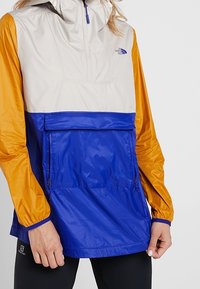 Lätt, färgblockerad vindjacka i beige, orange och blått. Har en halvdragkedja, ficka fram och elastiska ärmmuddar. Slät textur.