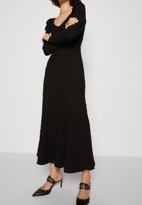 Svart långärmad klänning med ett texturerat mönster och fyrkantig halsringning. Bärs med svarta klackskor med spetsig tå som har gyllene cirkulära detaljer.