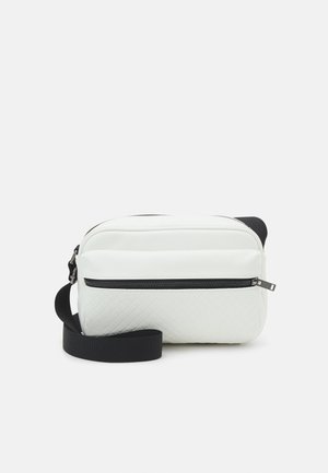 Sac à bandoulière blanc en matériau texturé, avec une poche zippée noire à l'avant, une bandoulière ajustable et un design lisse et arrondi.