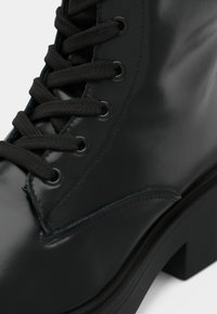 Μαύρη γυαλιστερή δερμάτινη μπότες μέχρι τον αστράγαλο με χοντρή σόλα και στρογγυλά μαύρα κορδόνια που περνάνε μέσα από μεταλλικές θηλιές.