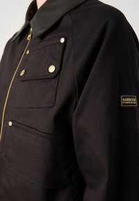 Jachetă din pânză neagră cu guler din piele maro, fermoar auriu, două buzunare frontale și un mic patch logo auriu pe mânecă.
