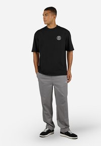 Svart t-skjorte med korte ermer og et sirkulært logo på brystet, kombinert med lysegrå bukser og svarte joggesko med hvite detaljer.