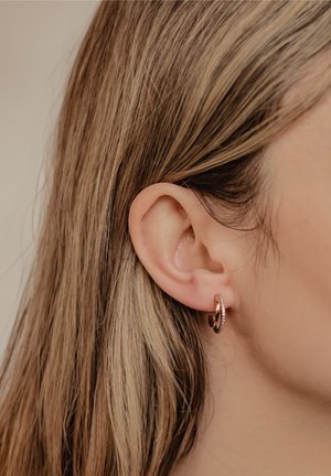 Ohrringe aus Roségold mit strukturierter Oberfläche und einem kleinen, klaren Akzentdetail, an einem Ohr eines Models mit glattem Haar im Hintergrund präsentiert.