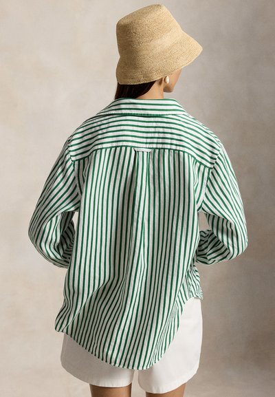 Žena v béžovom klobúku bucket, vo veľkej košeli s vertikálnymi zeleno-bielymi pruhmi a bielych krátkych nohaviciach, otočená chrbtom na jednobarevnom pozadí.