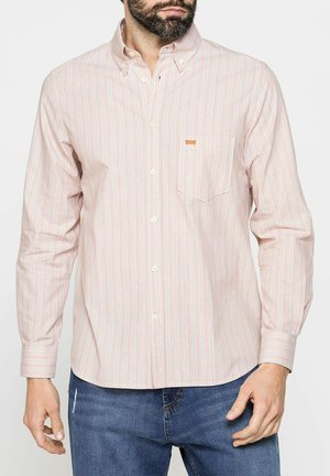 Uomo che indossa una camicia a righe pastello con taschino sul petto e jeans blu, in piedi con le mani rilassate lungo i fianchi.
