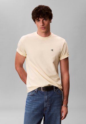 Ung mand iført en cremefarvet kortærmet t-shirt med oprullede ærmer, blå jeans, sort bælte og sølvarmbånd, stående foran en grå baggrund.