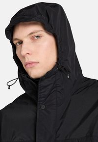 Mustan vedenpitävän takin, jossa on huppu, teksturoitu kangas, nyöritettävät suljintekniikat ja nappi kauluksessa säädettävää istuvuutta varten.