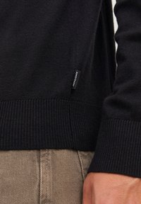 Czarny dzianinowy sweter z mankietami i dolnym brzegiem, z metką Jack & Jones, noszony na beżowych dżinsowych spodniach, ręka spoczywająca obok biodra.