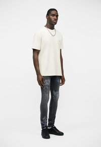 Mężczyzna w kremowej koszulce z krótkim rękawem, czarnym naszyjniku z łańcuszka, ciemnoszarych, wzorzystych obcisłych jeansach i czarnych sneakersach, stojący na białym tle.