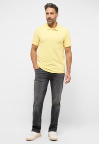 Sárga polo ing klasszikus gallérral, szürke farmernadrággal és fehér sportcipővel párosítva. Az anyag sima, fazonja pedig laza.