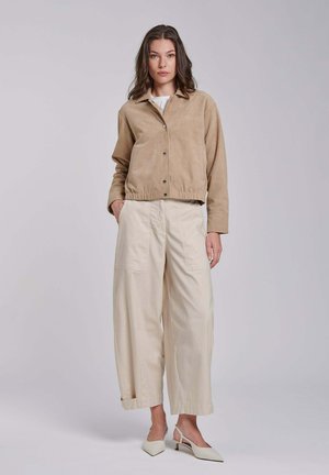 Vrouw staand, gekleed in een zandkleurige jas met knopen, brede beige broek met zakken en witte puntige slingback-hakken tegen een neutrale achtergrond.