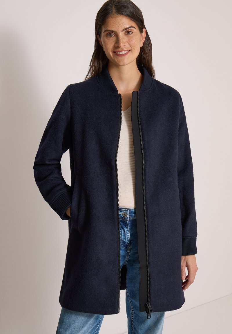 Femme aux cheveux foncés portant un manteau bleu marine zippé par-dessus un haut blanc et un jean bleu, souriante, une main dans la poche.