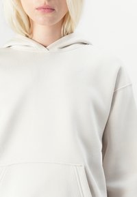 Light beige hoodie with a kangaroo pocket, soft texture, adjustable hood, and a subtle embossed logo on the chest.