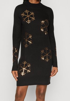 Robe-pull noire à manches longues avec des motifs flocon de neige en sequins dorés sur le devant, les manches et l'ourlet, portée par une personne debout.