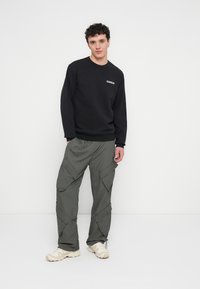 Felpa nera con un piccolo logo bianco sul petto, abbinata a pantaloni cargo verde oliva ampi, caratterizzati da multiple tasche e coulisse.