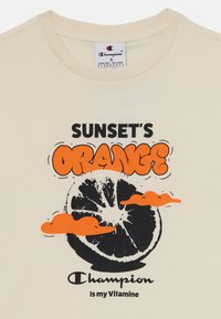 Grietinė t-shirt su grafiniu dizainu, vaizduojančiu juodą apelsino pusę, oranžines debesis ir tekstą "SAULĖS NUSILEIDIMO ORANŽINIS" juoda ir oranžine spalvomis.