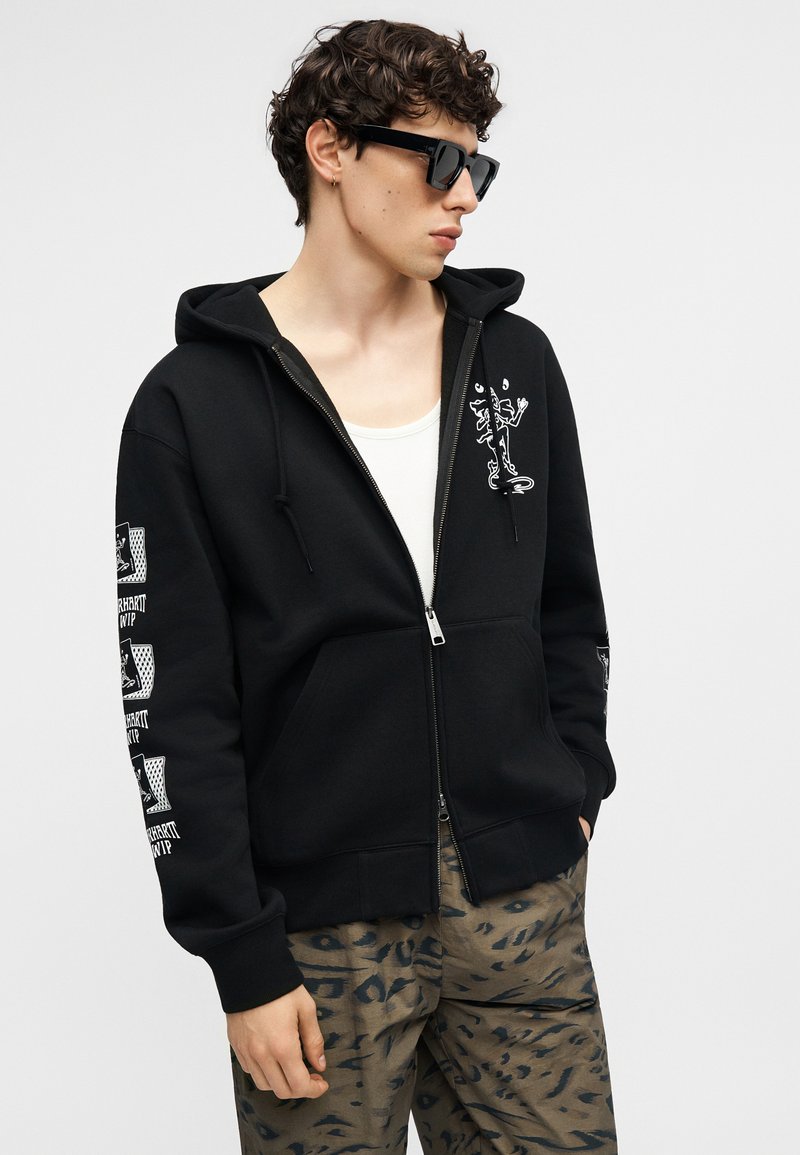 Junger Mann, der eine schwarze Kapuzenjacke mit Reißverschluss und weißem Grafikmotiv, Tarnhosen, ein weißes Tanktop und eine schwarze Sonnenbrille trägt, mit den Händen in den Taschen stehend.