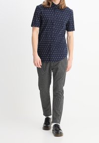 Camicetta blu navy a maniche corte con motivo geometrico bianco, abbinata a pantaloni slim-fit a scacchi bianchi e neri e scarpe nere.