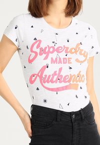 Camiseta de algodón blanca con mangas cortas, que presenta un diseño gráfico en texto en rosa y naranja que dice "Superdry MADE Authentic" y pequeños patrones geométricos dispersos.