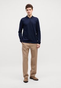 Camisa polo de zip azul-marinho de mangas longas, com tecido texturizado, combinada com calças bege e botas de camurça castanhas.