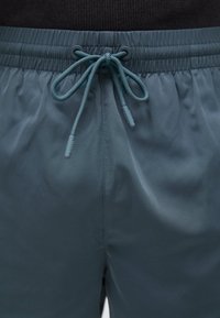 Costume da bagno in rete color verde scuro, con vita elastica e chiusura con cordino. Texture liscia con una leggera lucentezza.