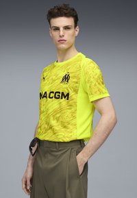 Żółta sportowa koszulka z abstrakcyjnym wzorem, czarnym logo i napisem, krótki rękaw, okrągły dekolt, lekki materiał, w komplecie z oliwkowymi spodniami.