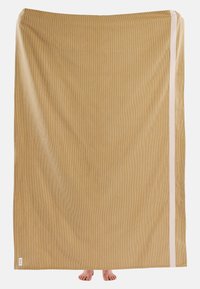 Velká béžová tkanina s texturovaným povrchem, s bílým pruhem podél jednoho okraje. Ve spodní části jsou vidět dvě bosé nohy.