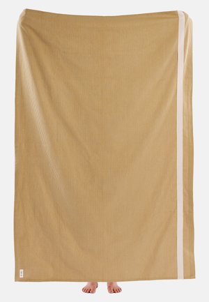 Veliko bež tkanino z teksturirano površino, ob strani pa bela črta. Na dnu so vidne dve bose nogi.