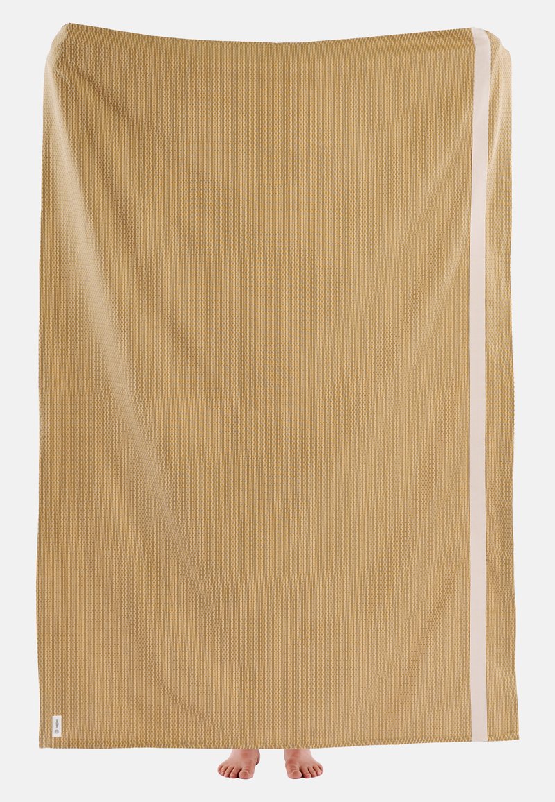 Velká béžová tkanina s texturovaným povrchem, s bílým pruhem podél jednoho okraje. Ve spodní části jsou vidět dvě bosé nohy.