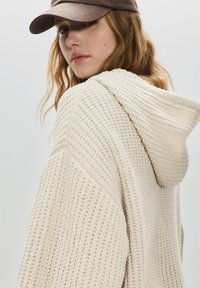 Hanorac crem tricotat cu un design texturat și glugă. Purtați cu o șapcă maro, evidențiind modelul moale și gros de tricotaj.