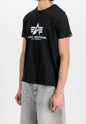 Osoba nosząca czarną koszulkę Alpha Industries z białym logo i napisem, w połączeniu z jasnoszarymi wyblakłymi dżinsami, pokazująca tatuaże na ramionach i pierścionki.