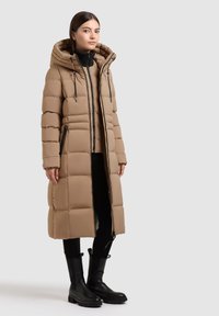 Beige színű puffertakaró, kapucnival, steppelt kialakítással, elülső cipzárral, oldalsó zsebekkel és hosszú sziluettel, fekete bokacsizmákkal párosítva.