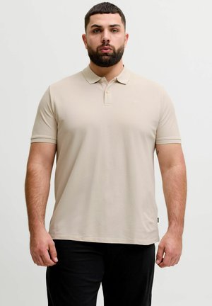 Man with short dark hair and beard wearing a beige polo shirt and black pants, standing against a plain light background.
