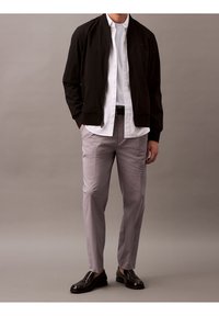 Schwarze Bomberjacke über einem weißen Hemd mit Knöpfen, kombiniert mit hellgrauen Hosen und schwarzen Loafers. Einfaches Design mit glatter Textur.