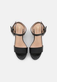 Svarta klacksandaler med ett slätt övre rem och en krokodilpräglad nedre rem. Spänne vid vristen, beige innersula med logotyp.